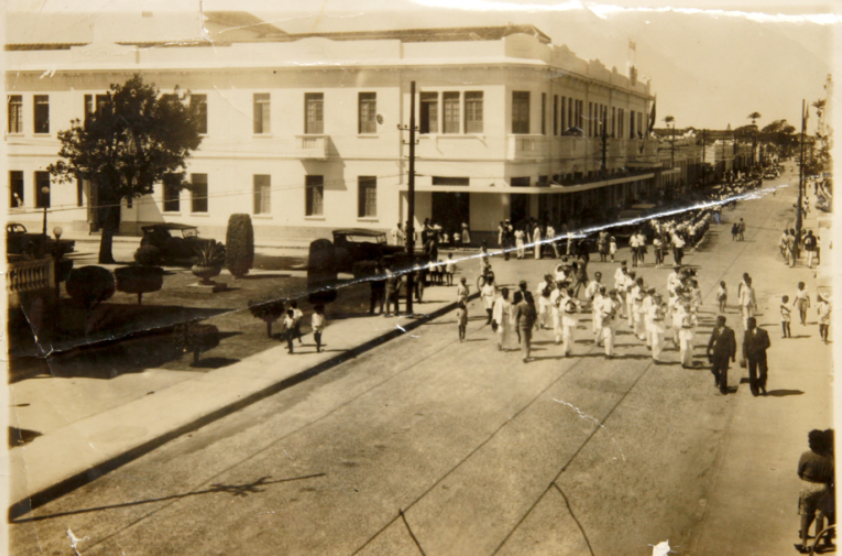 Fotografia antiga, em tom sépia, mostrando uma rua movimentada no centro de Macaé. Um prédio grande de dois andares ocupa o lado esquerdo da imagem, enquanto uma banda ou grupo musical desfila no meio da rua, acompanhada por moradores e transeuntes. A cena registra um momento histórico da cidade, com pessoas observando o cortejo e carros antigos estacionados ao fundo.